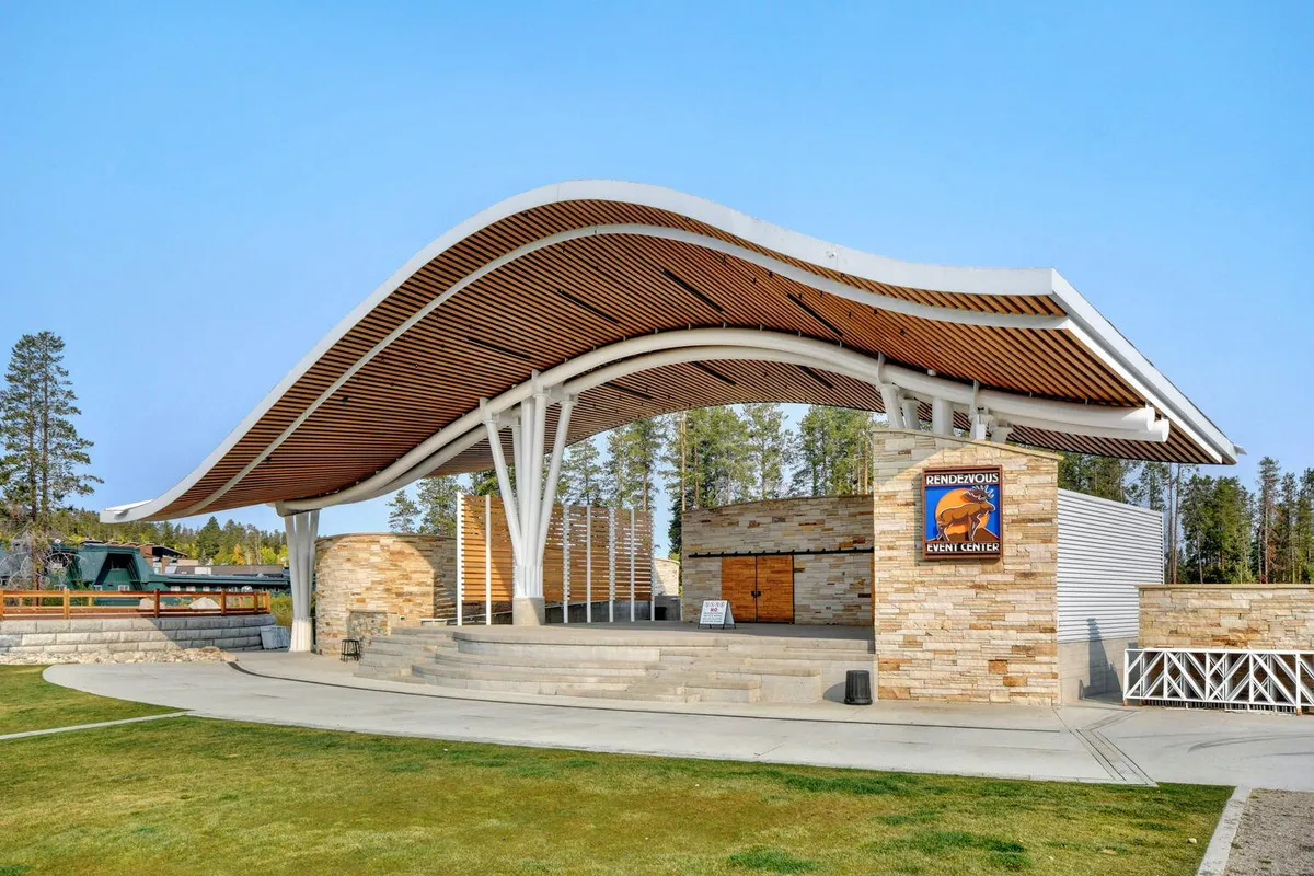 A lively exterior shot of a curved roof structure, stone steps, and community event center signage. The area is designed for gatherings, concerts, and outdoor activities, set against the scenic backdrop of the Rockies. The openness hints at ample outdoor space for walking and play.
