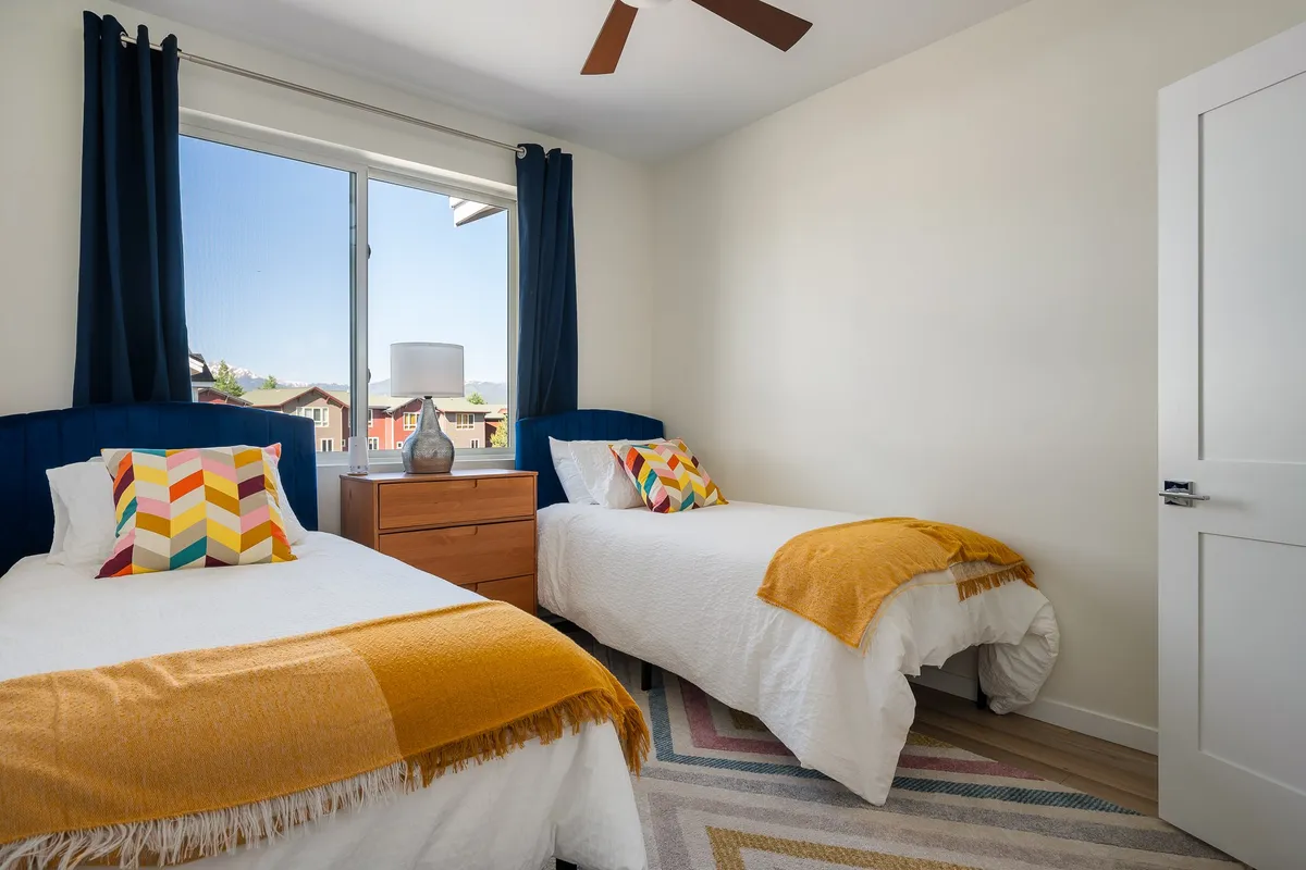 A cheerful twin bedroom featuring two twin beds with colorful throw pillows, a wooden nightstand, and a large window that allows abundant daylight to fill the space. The setup is playful yet comfortable, ideal for children or friends.