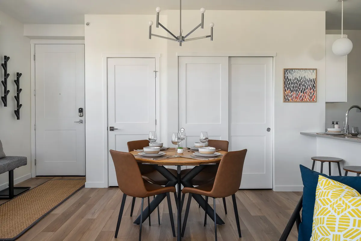 A welcoming dining area featuring a table set for four beneath a modern chandelier, with wall art and a partial view of the adjacent kitchen. The setting is bright and comfortable, inviting guests to share meals or conversation.