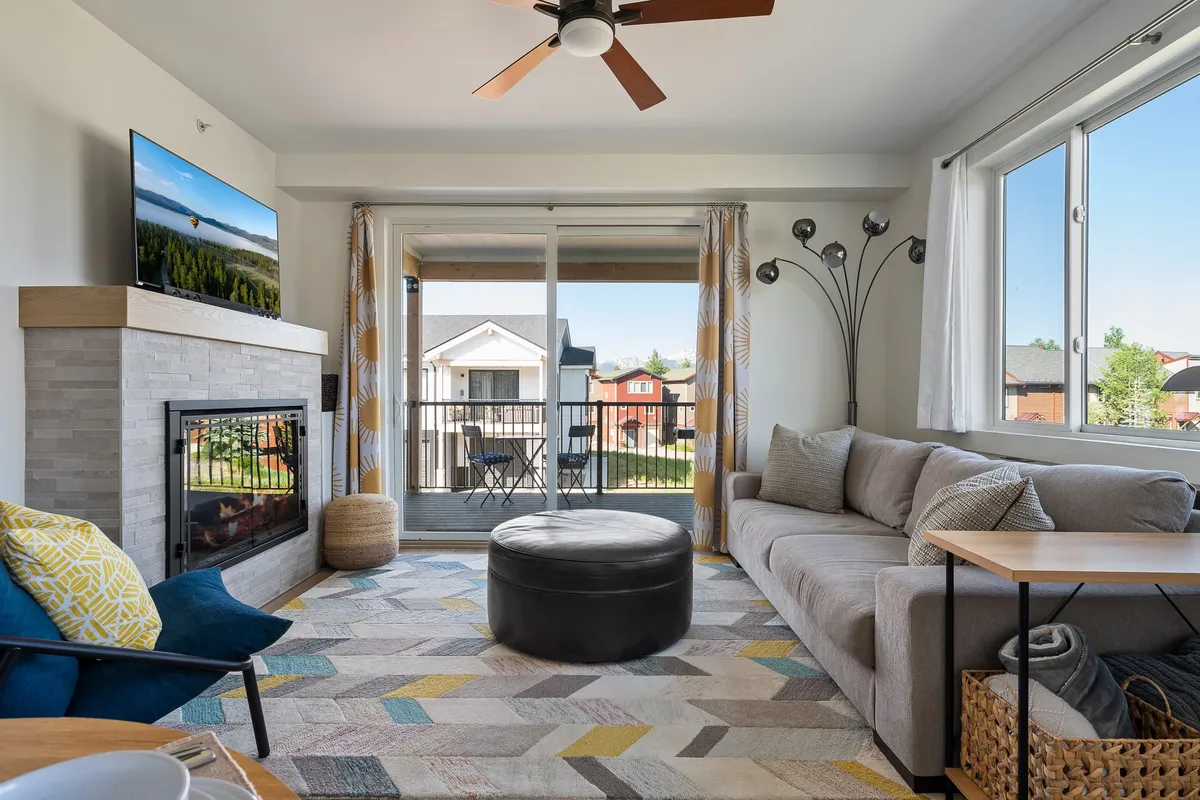 A cozy living area with a plush sofa, a color-changing electric fireplace, large coffee table, and a 65" HD streaming TV. The space extends to a balcony view, allowing natural light to fill the room. The setting is ideal for relaxation, movie nights, or board games by the fire.