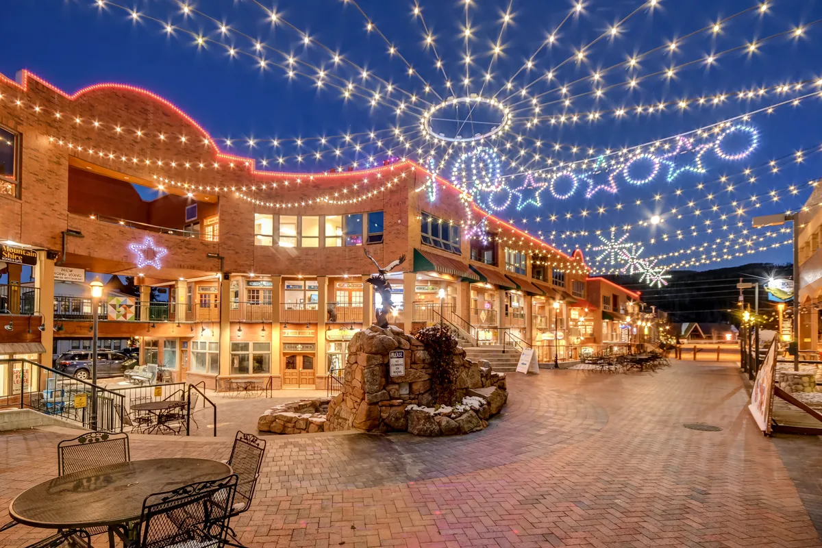 A lively exterior scene with decorative holiday lights, attractive building architecture, a paved walkway, and a statue centerpiece. The area buzzes with activity, featuring outdoor dining, shopping, and social gatherings—emphasizing the vibrant community atmosphere of the resort base area.