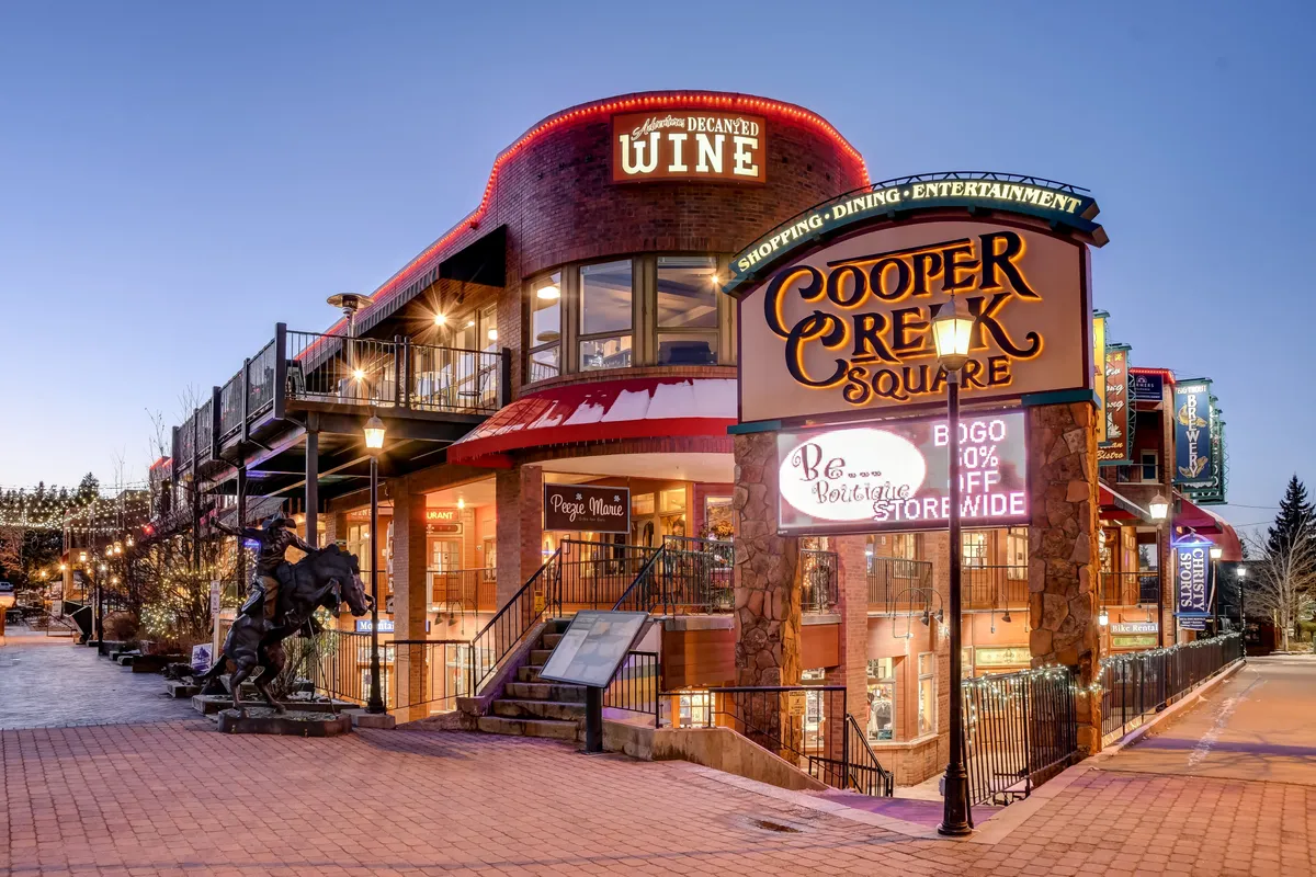 A bustling exterior shot showing the Cooper Creek Square sign, wine shop signage, and a staircase leading up. The area is lively with hints of shopping, dining, and year-round community happenings, inviting exploration for guests and their dogs alike.