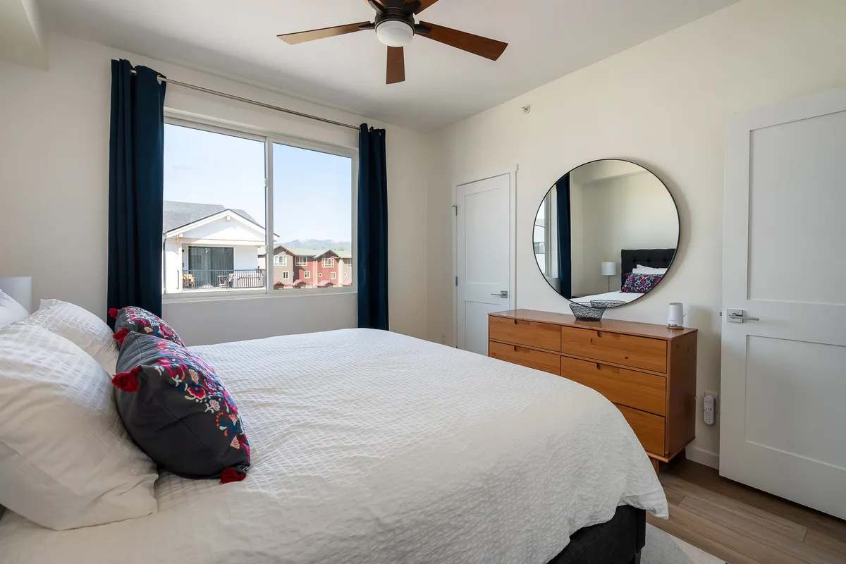 This image shows a serene bedroom with a large bed dressed in white linens, a circular mirror above a wooden dresser, and a window revealing natural light and scenic views. The overall atmosphere is calm, cozy, and inviting—ideal for relaxation and restful nights.
