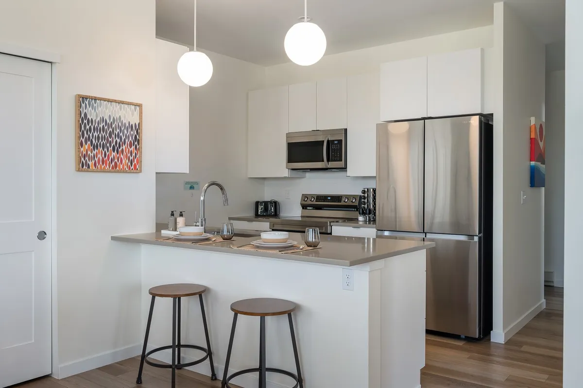 A bright, modern kitchen featuring a spacious quartz-topped island, gleaming stainless steel appliances, chic pendant lights, and contemporary artwork on the wall. The island offers ample seating and workspace, perfect for meal prep, casual dining, or gathering with friends and family.
