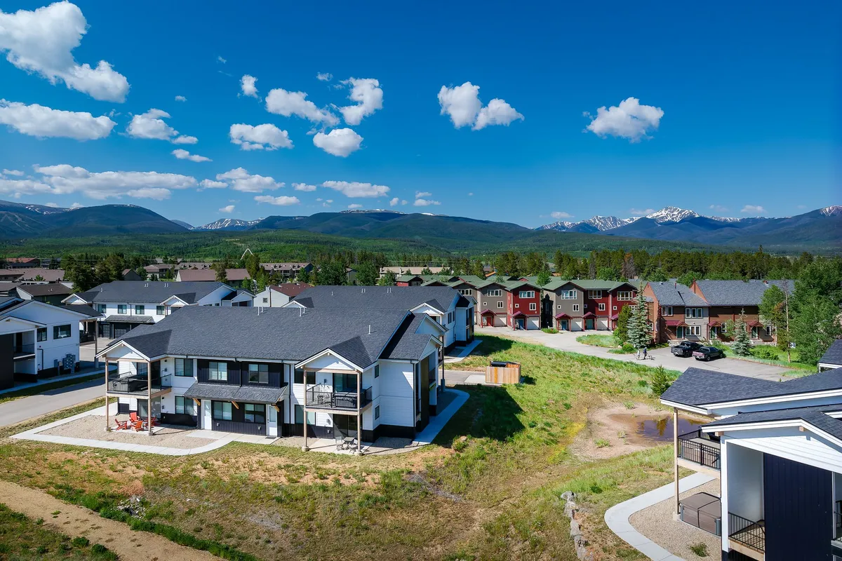 A welcoming exterior shot of modern apartment buildings set against a backdrop of mountains, with families and neighbors enjoying picnics, nature walks, and outdoor gatherings in well-maintained green spaces. The image emphasizes community, family enjoyment, and the stunning natural surroundings.