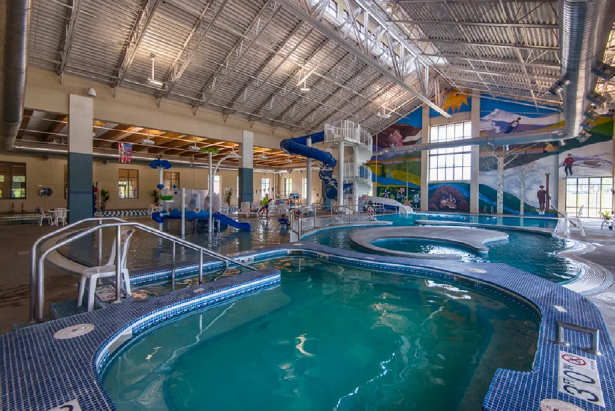 A lively indoor rec center scene with families and children enjoying water slides, swimming pools, and a vibrant mural. The atmosphere is energetic and joyful, highlighting recreational amenities available to guests regardless of the season.