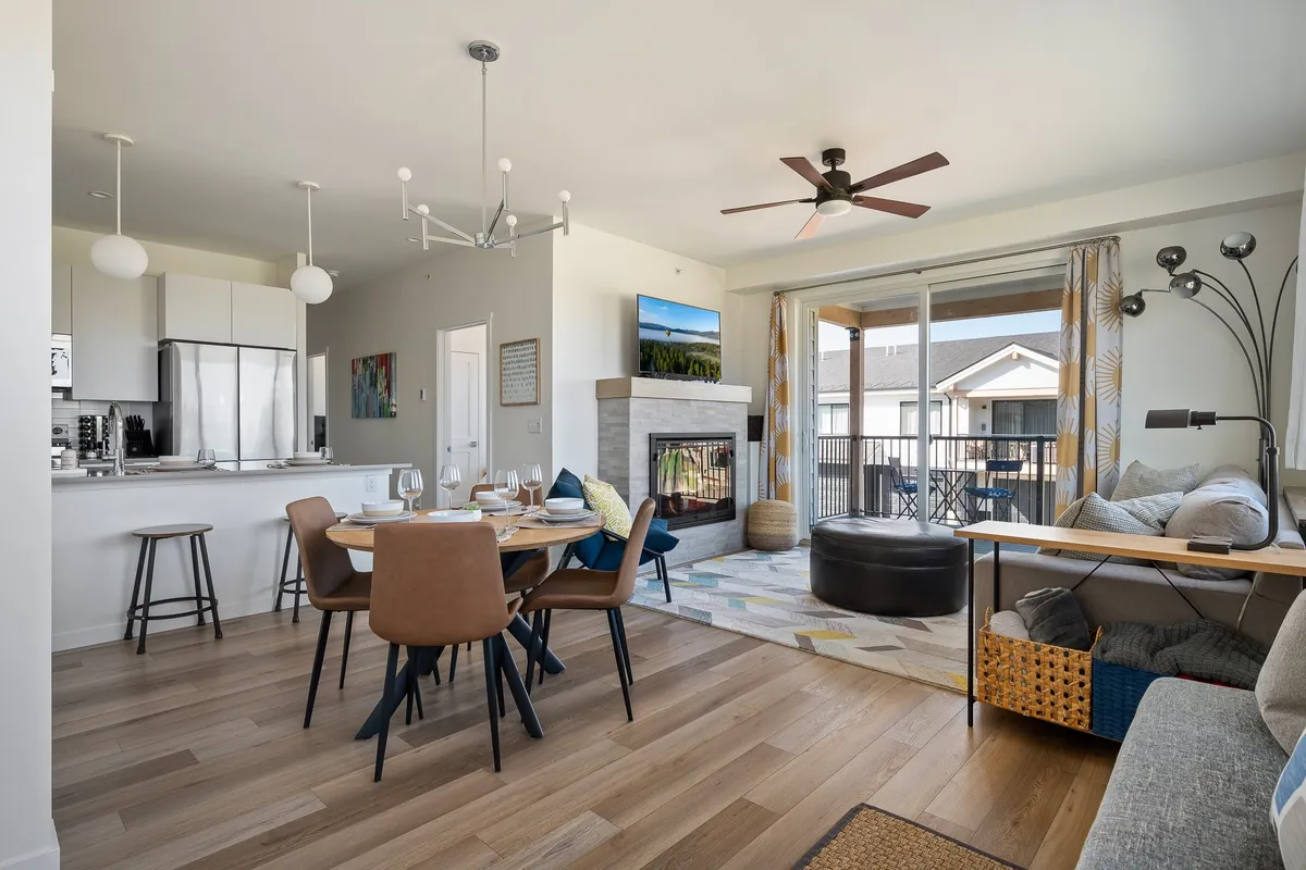 The image showcases a bright and inviting open-concept living area featuring a stylish dining table set for a meal, a cozy seating area oriented around a modern fireplace with a TV above, and expansive windows revealing scenic mountain views. The overall ambiance is warm and welcoming, suggesting a space perfect for gathering and relaxation.